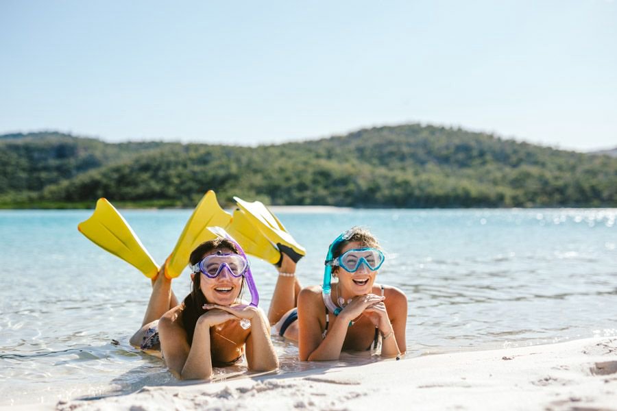 Two women wearing snorkel masks and yellow fins lie on shallow white sand near calm water, smiling at the camera in the Whitsundays