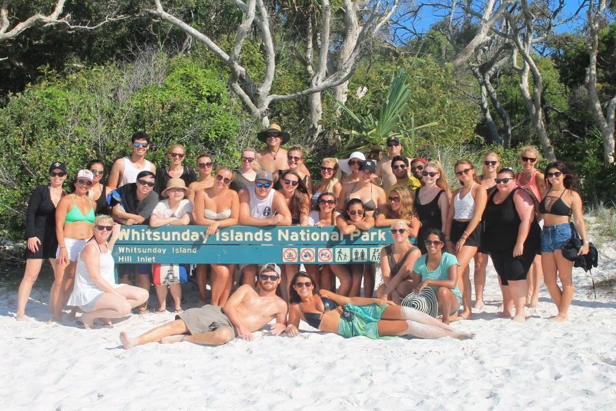 Large group of travellers posing together on white sand in front of the Whitsunday Islands National Park sign at Hill Inlet, surrounded by tropical bushland