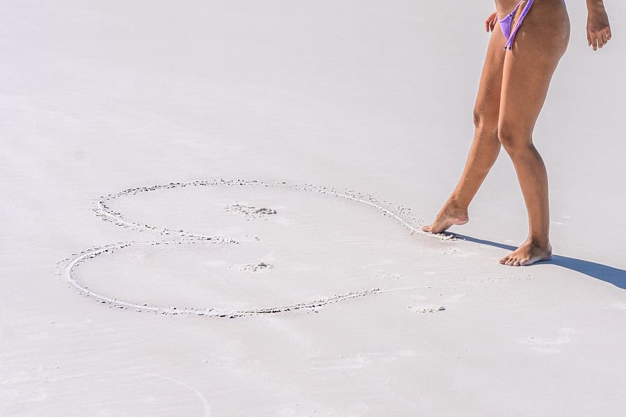 A woman walks barefoot along Whitehaven Beach beside a large heart shape drawn in the soft white sand