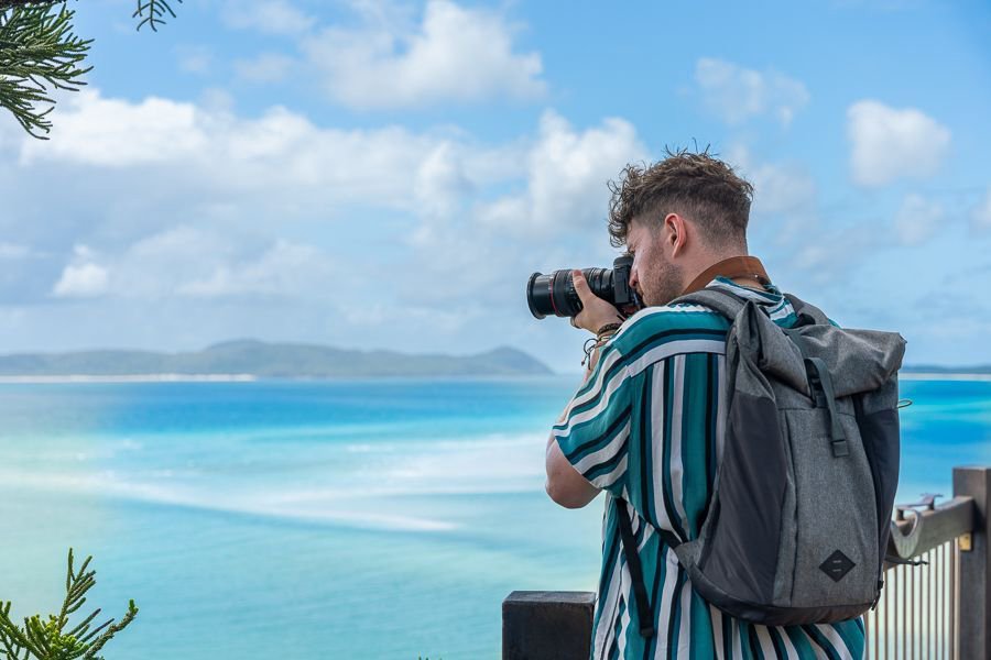A man with a backpack taking a photo of the turquoise waters and swirling sands of Whitehaven Beach from Hill Inlet Lookout in the Whitsundays