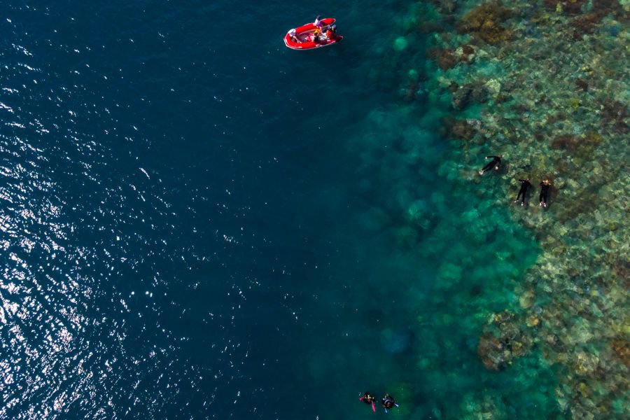 Aerial view of snorkellers exploring a vibrant coral reef in clear turquoise water, with a red inflatable boat floating nearby