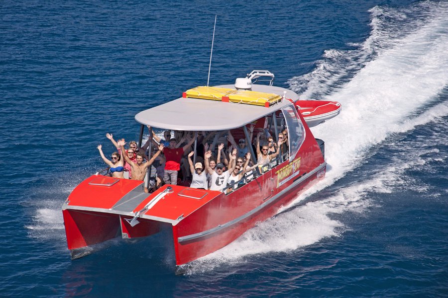 A red ThunderCat speedboat cruises across deep blue ocean waters in the Whitsundays, with passengers onboard smiling and waving as the boat leaves a white wake behind