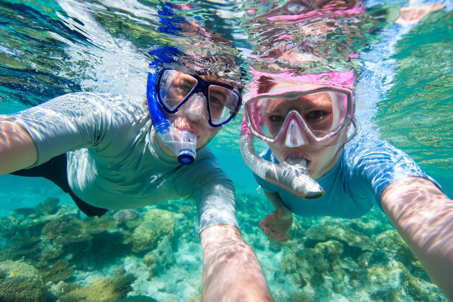 Two snorkellers wearing blue and pink masks take an underwater selfie above vibrant coral reef in clear turquoise water