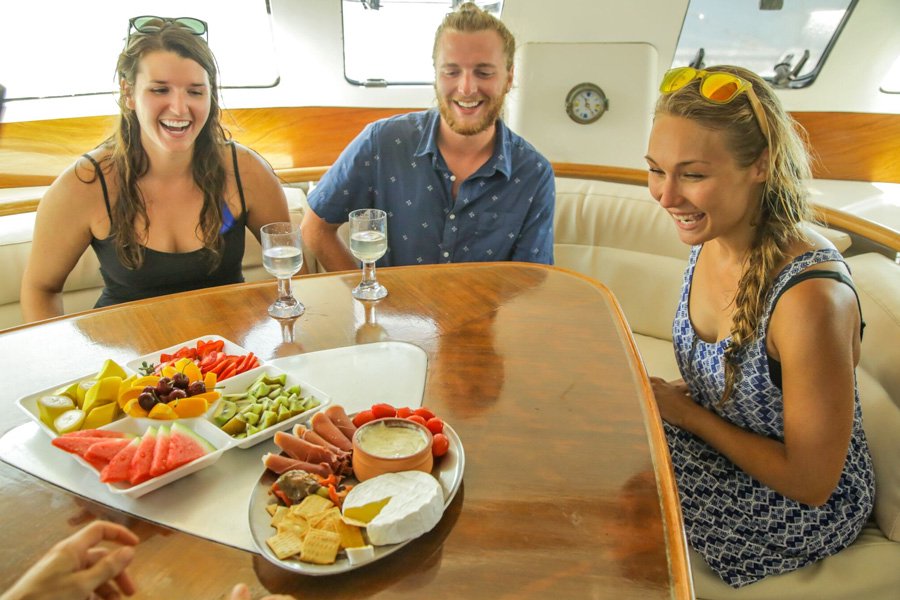 Three friends sitting around a wooden table inside a yacht saloon, smiling at a fresh fruit and cheese platter with drinks in hand