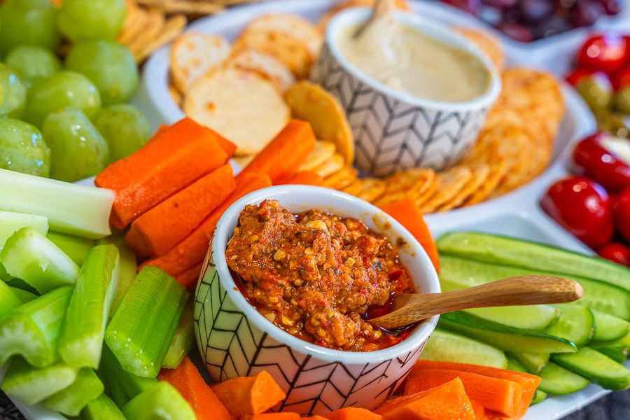 Close up of a colourful grazing platter featuring fresh celery, carrot sticks, cucumber slices, cherry tomatoes, grapes, crackers and two dipping sauces