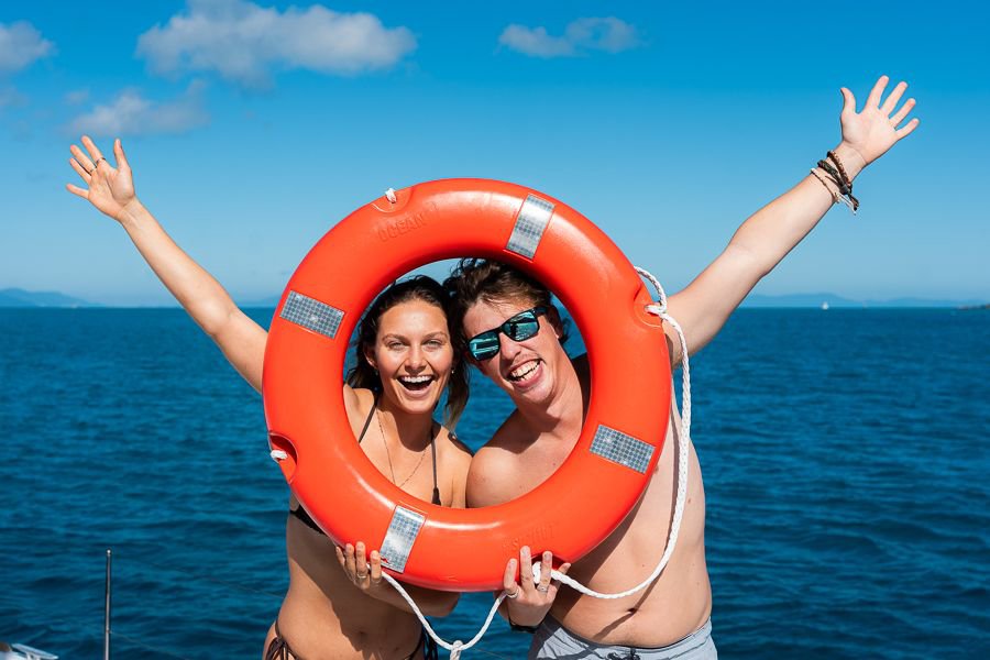 Two friends smiling with their faces framed inside a bright orange lifebuoy on a boat, with blue ocean and clear sky in the background