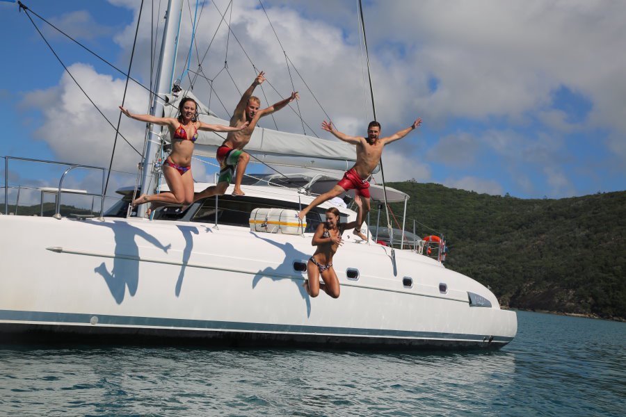 Four friends in swimwear leap from the side of a white catamaran into clear blue water, with a lush green island in the background