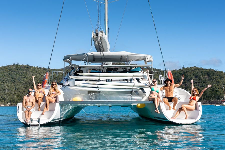 Group of friends sitting on the back of a white catamaran in turquoise Whitsundays waters, smiling and raising their arms on a sunny day
