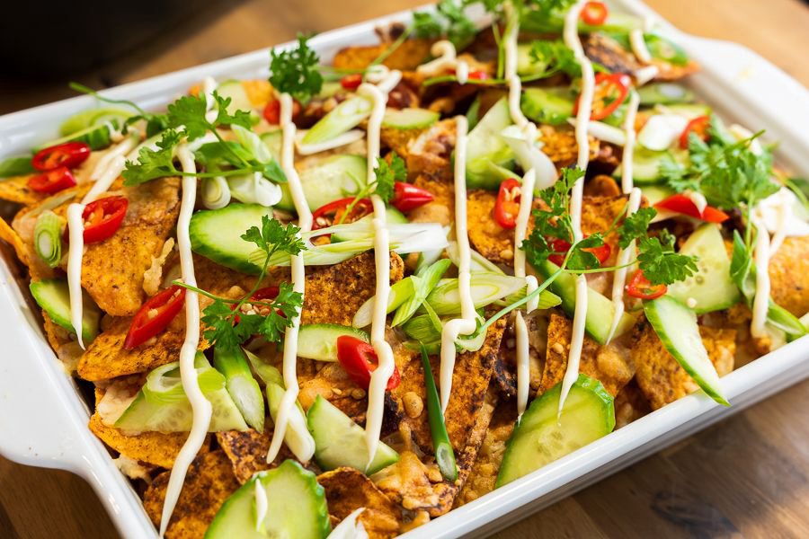 A platter of gourmet nachos topped with grilled chicken, cucumber, sliced chilli, fresh herbs and drizzled sauce, served on a boat