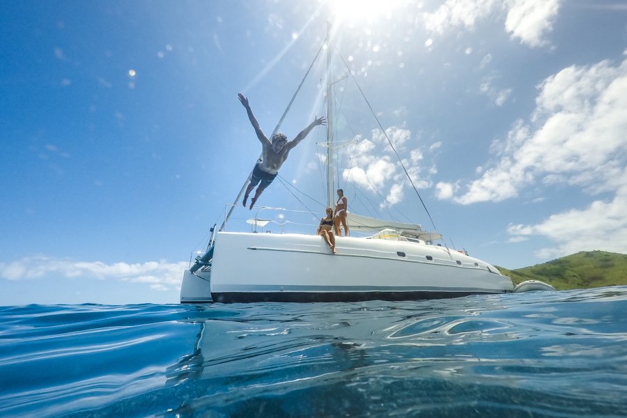 A man dives off the front of a white catamaran into clear blue water while two women watch from the deck, with bright sunshine and a lush island in the background