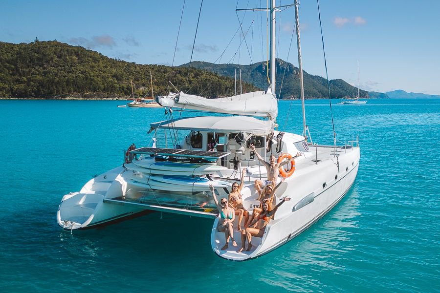 Group of friends relaxing and posing on the front deck of a white catamaran anchored in bright turquoise waters in the Whitsundays, with lush green islands in the background