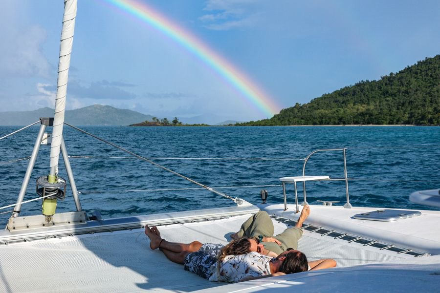 Couple lying on the deck of a catamaran while a bright rainbow arches over turquoise water and lush green islands in the Whitsundays