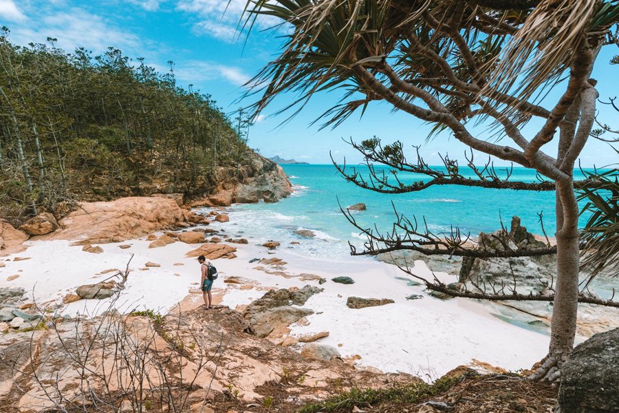 Traveller standing on rocky shoreline overlooking a secluded white sand beach with turquoise water in the Whitsundays