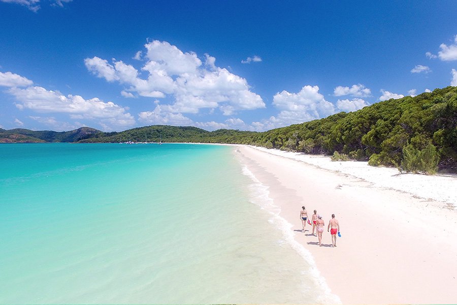 Aerial view of three people walking along a quiet white sand beach with turquoise water and lush green hills in the Whitsundays