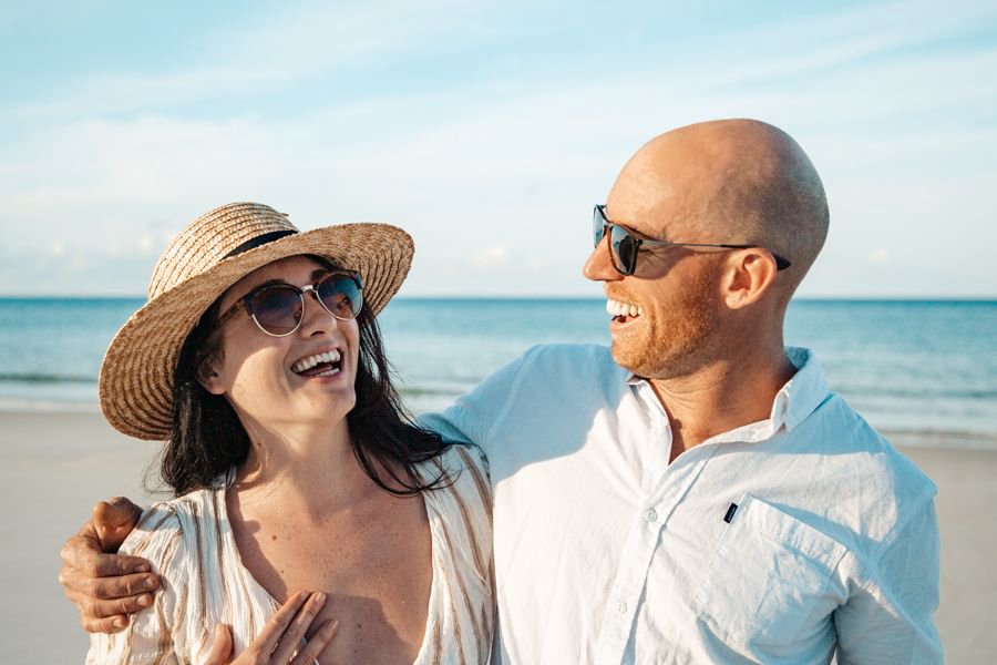 A smiling couple wearing sunglasses stands close together on a sunny beach, laughing with the ocean in the background