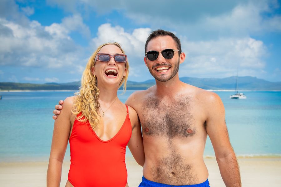 A smiling couple in swimwear standing on the sandy shore of Whitehaven Beach with turquoise water and a sailboat in the background