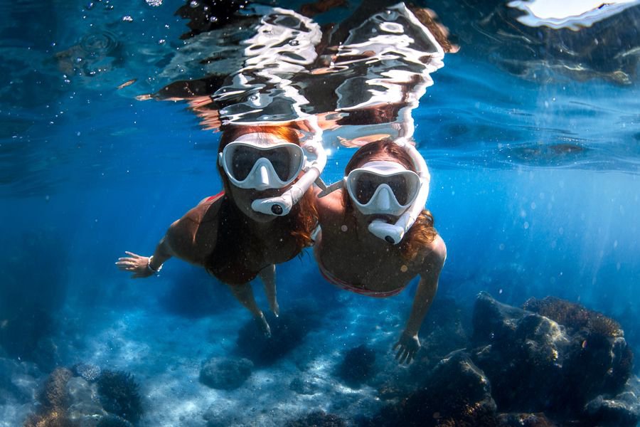 Two women snorkelling underwater with full-face masks above coral reef in clear blue water in the Whitsundays