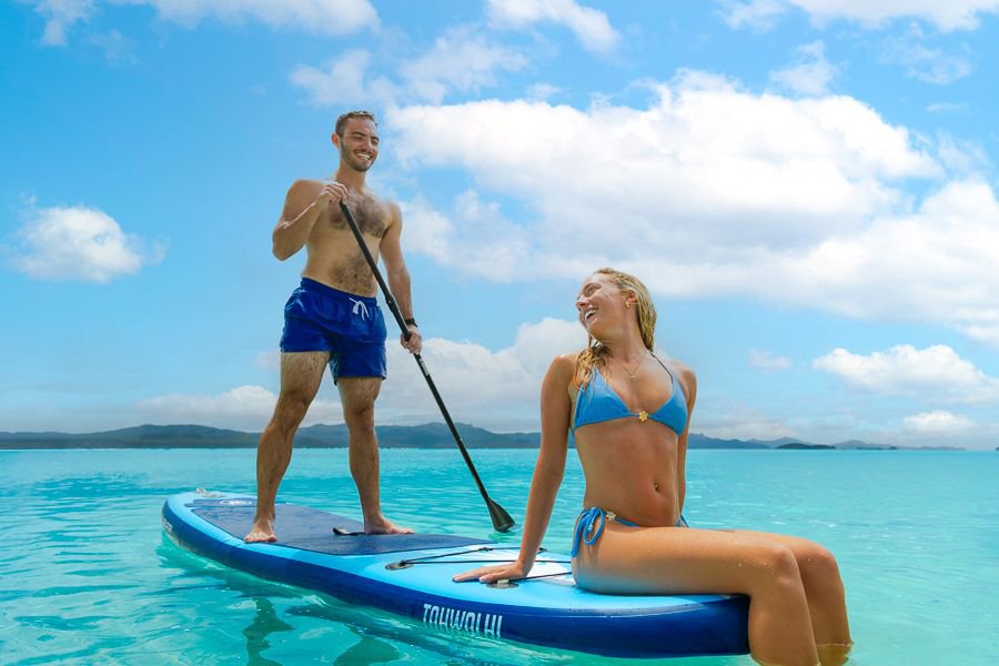 A couple enjoying stand-up paddleboarding on calm turquoise waters in the Whitsundays, with the man paddling while the woman sits on the board under a bright blue sky