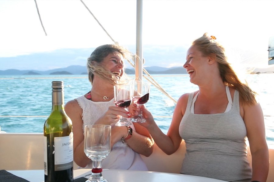 Two women laughing and clinking glasses of red wine while relaxing on a yacht at sunset, with ocean and island views in the background