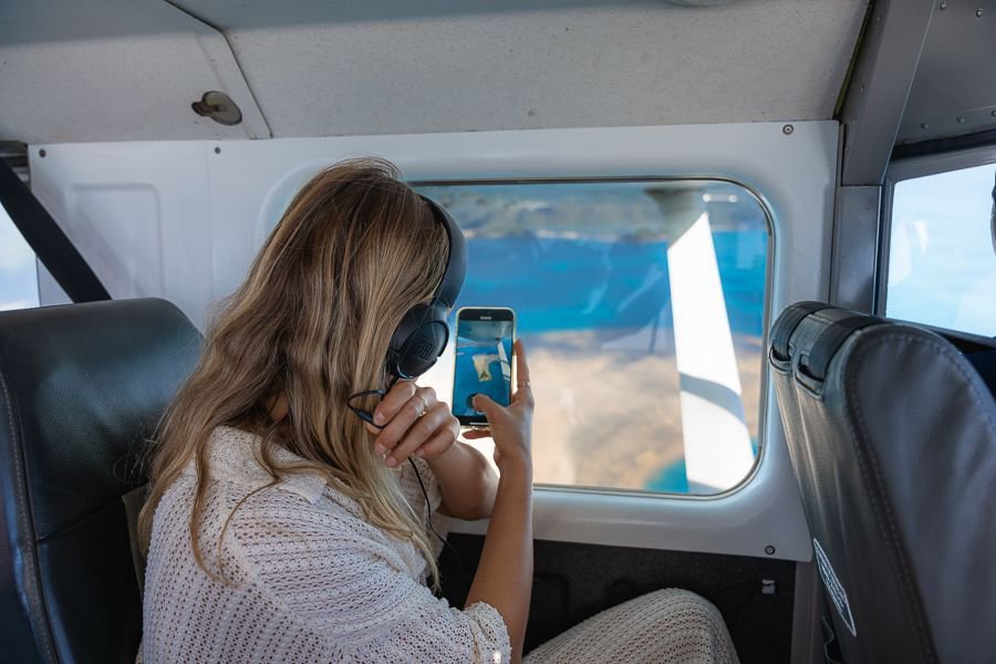 Passenger wearing a headset inside a small aircraft taking a photo through the window of turquoise reef waters below.