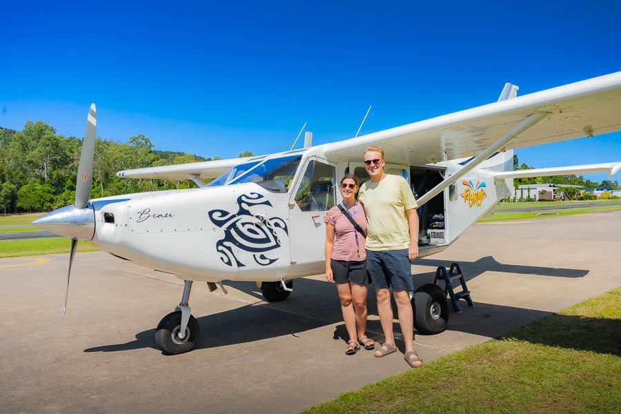 A smiling couple stands in front of a small white scenic flight plane on the runway, ready for a Whitsundays aerial tour.