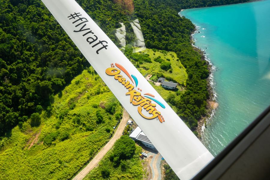 View from a scenic flight plane wing branded Ocean Rafting flying over lush green coastline and turquoise waters in the Whitsundays.