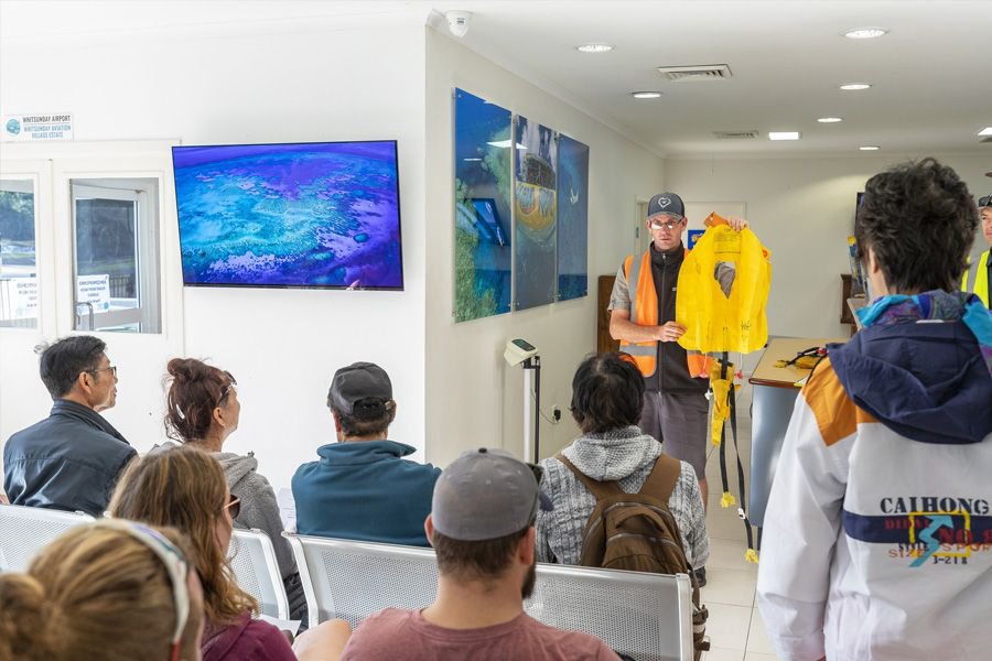 Group of passengers seated inside the Whitsunday Airport terminal listening to a staff member in a high-visibility vest giving a pre-flight safety briefing.