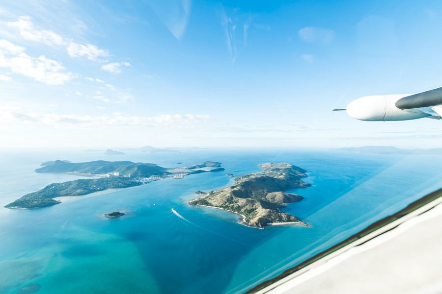 Aerial view from a small aircraft window over the Whitsunday Islands, showing lush green islands surrounded by turquoise ocean and reef formations, with part of the plane’s wing visible.