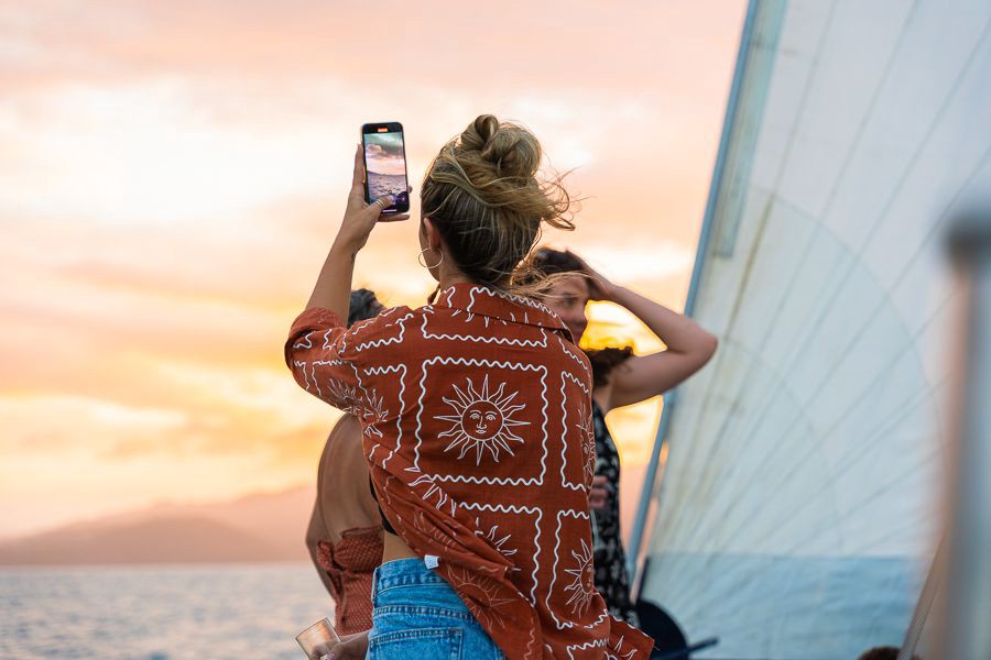 Woman taking a photo of a vibrant sunset on her phone while standing on a sailing boat in the Whitsundays, Queensland.