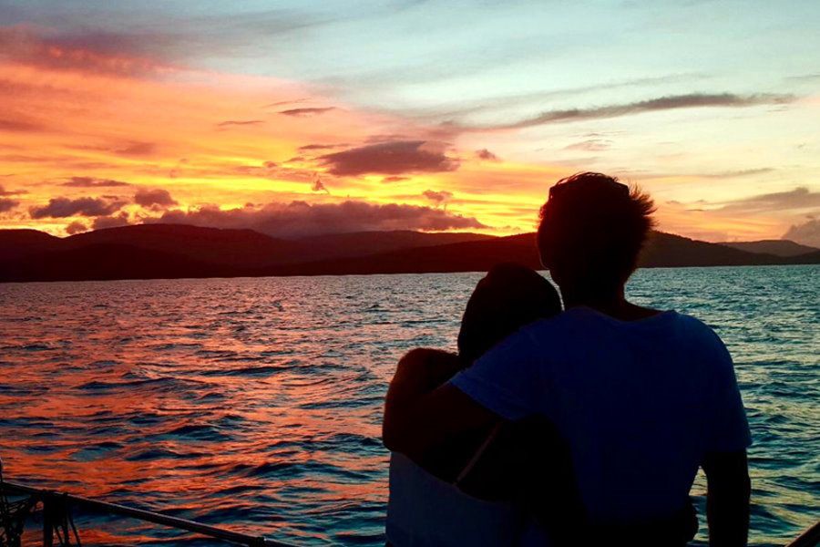 Silhouetted couple embracing on a sailing boat while watching a vibrant red and orange sunset over the Whitsunday Islands