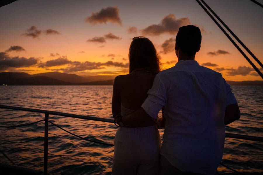 Couple embracing on the deck of a sailing boat while watching a vibrant orange sunset over the Whitsunday Islands in Queensland, Australia.