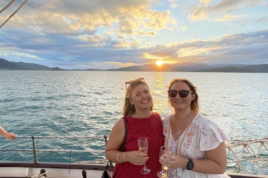 Two women holding champagne glasses on a sailing boat at sunset with calm ocean waters and island mountains in the Whitsundays, Queensland.