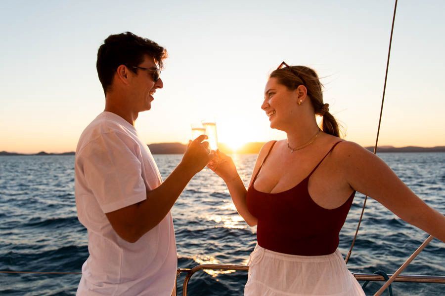 Couple toasting drinks on the deck of a sailing boat at sunset with the ocean and island silhouettes in the background in the Whitsundays, Queensland.