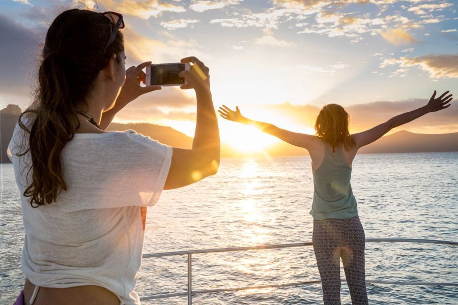 Woman taking a photo of her friend with arms outstretched while watching a golden sunset from a sailing boat in the Whitsunday Islands, Queensland.