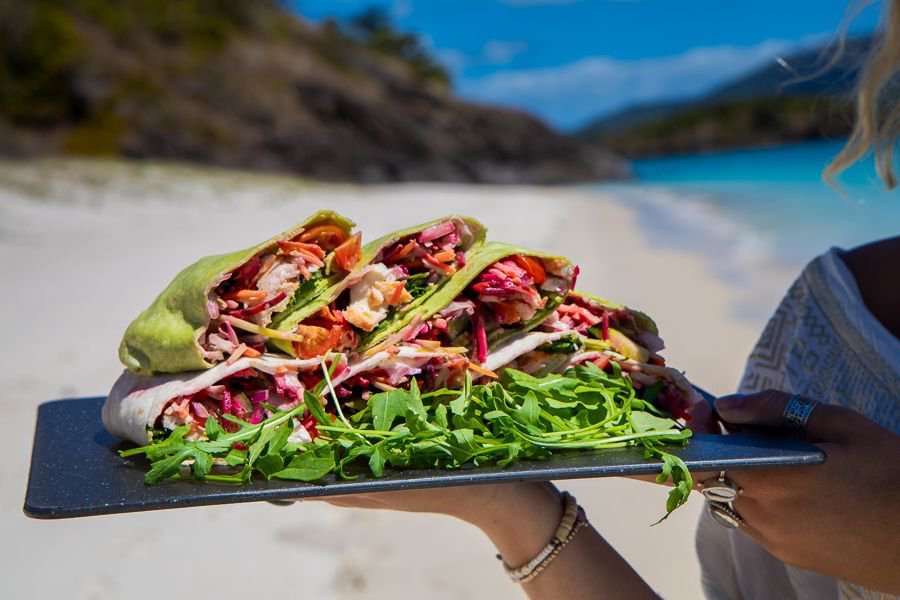 Close-up of freshly made gourmet wraps filled with vegetables and meat, served on a black platter with arugula, with a white sandy beach and turquoise ocean in the background.