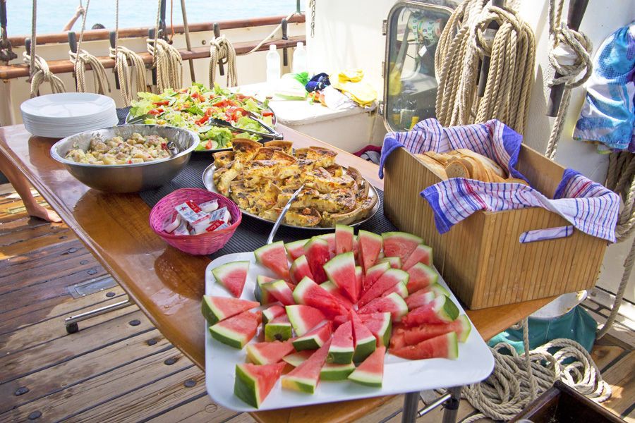 A wooden table set on the deck of a sailing boat with fresh plated food—including salads, seafood, bread and drinks—against a backdrop of blue ocean and sky.