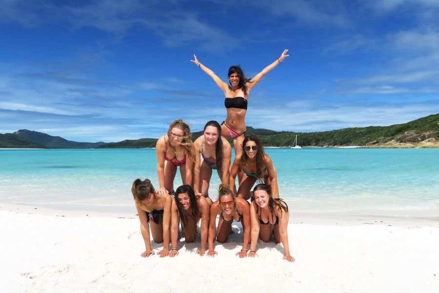 Group of women in bikinis forming a playful human pyramid on the white sand of Whitehaven Beach with turquoise water and green hills behind them.