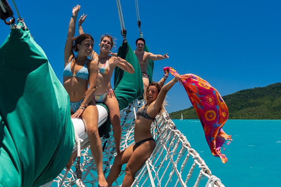 Four friends sitting and laughing together on the bow net of a sailing catamaran, with turquoise water and island hills in the background.