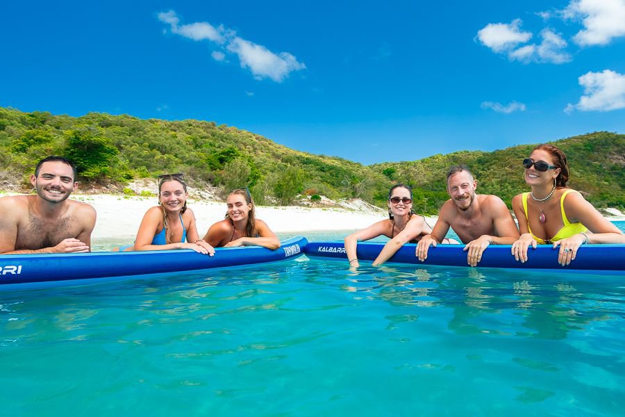 Group of friends smiling and leaning on paddleboards in clear turquoise water surrounded by lush green islands in the Whitsundays.