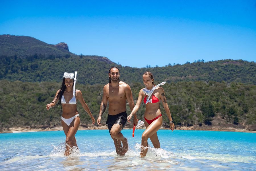 Three friends in swimwear walking through shallow turquoise water with a lush island backdrop in the Whitsundays.