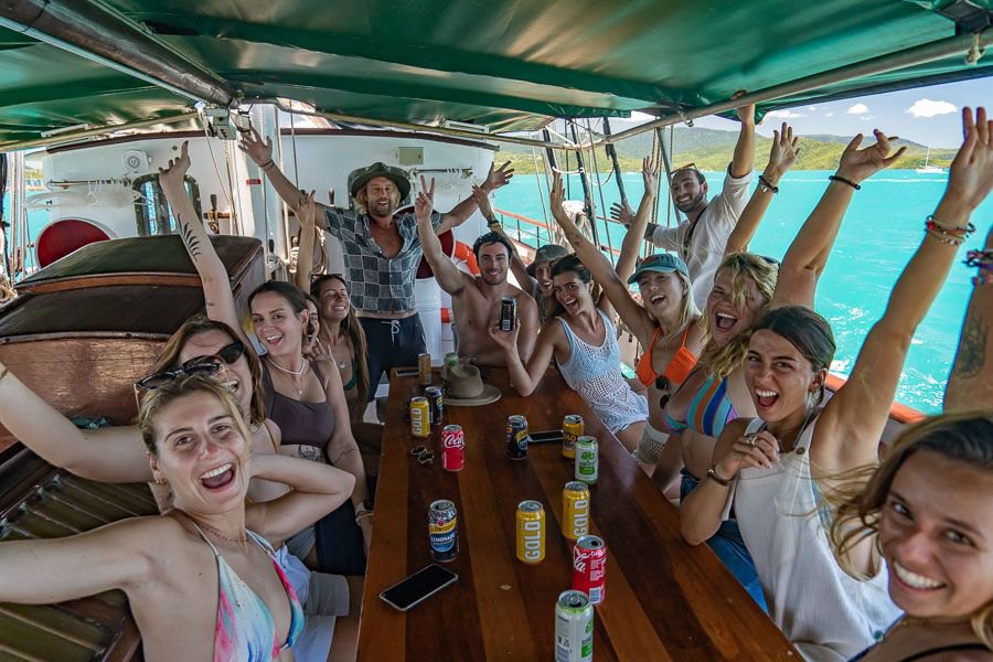 A lively group of friends cheering with their hands in the air around a table on a sailing boat, surrounded by bright turquoise ocean.