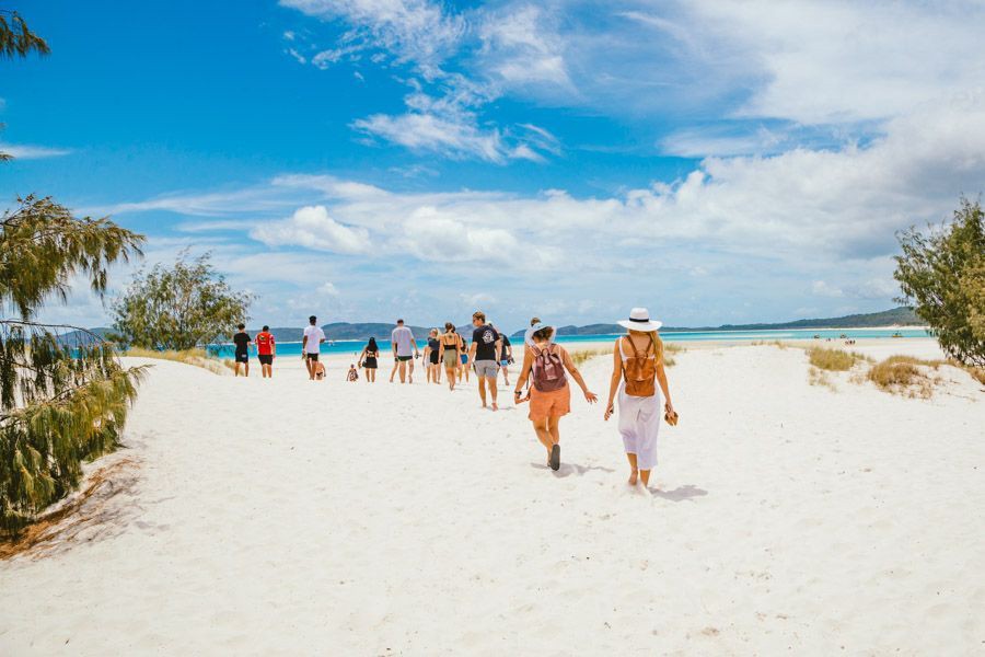 A group of people walking across bright white silica sand toward the turquoise waters and islands of Hill Inlet and Whitehaven Beach in the Whitsunday Islands under a partly cloudy blue sky.