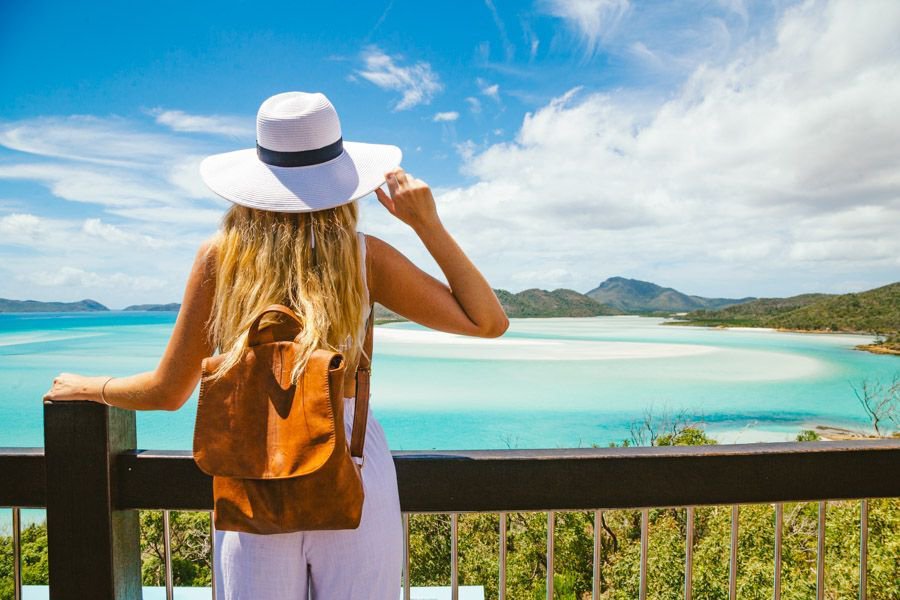 Woman wearing a white hat and backpack admiring the swirling white sands and turquoise waters of Hill Inlet from a lookout in the Whitsundays.