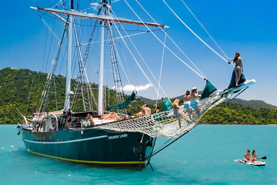 Guests lounging on the bow nets of a classic tall ship sailing in turquoise Whitsundays waters with lush islands in the background.