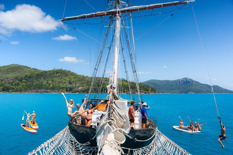 Group of friends relaxing and celebrating on the deck and bow net of a sailing boat in the Whitsundays, with paddleboarders floating nearby in clear turquoise water.