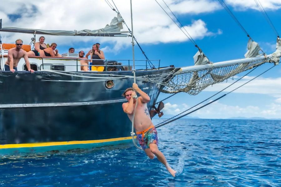 Guest swinging from a rope off the bow of a traditional tall ship into clear blue ocean waters, with friends watching and cheering from the deck above.