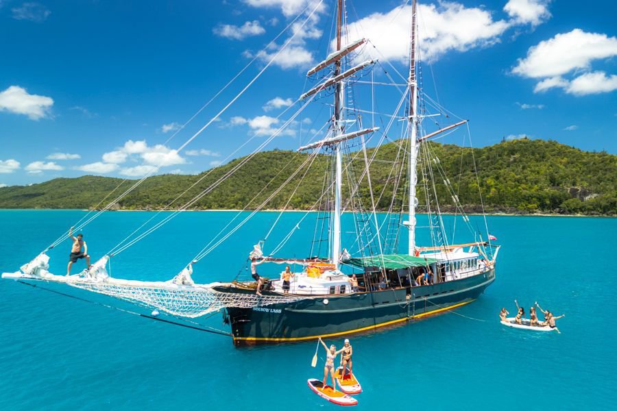 Classic tall ship anchored in turquoise Whitsundays waters with guests relaxing on deck and swimming nearby.