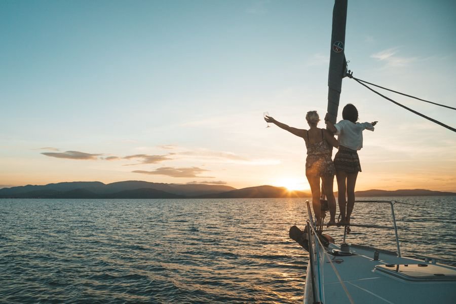 Two women standing at the bow of a sailing yacht at sunset in the Whitsundays, overlooking calm ocean and island silhouettes