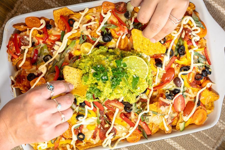 hands grabbing chips from a plate of nachos with colourful toppings