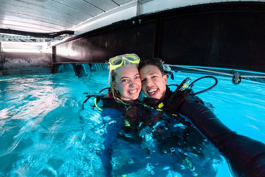 couple with scuba gear smiling under a boat while treading water over a coral reef